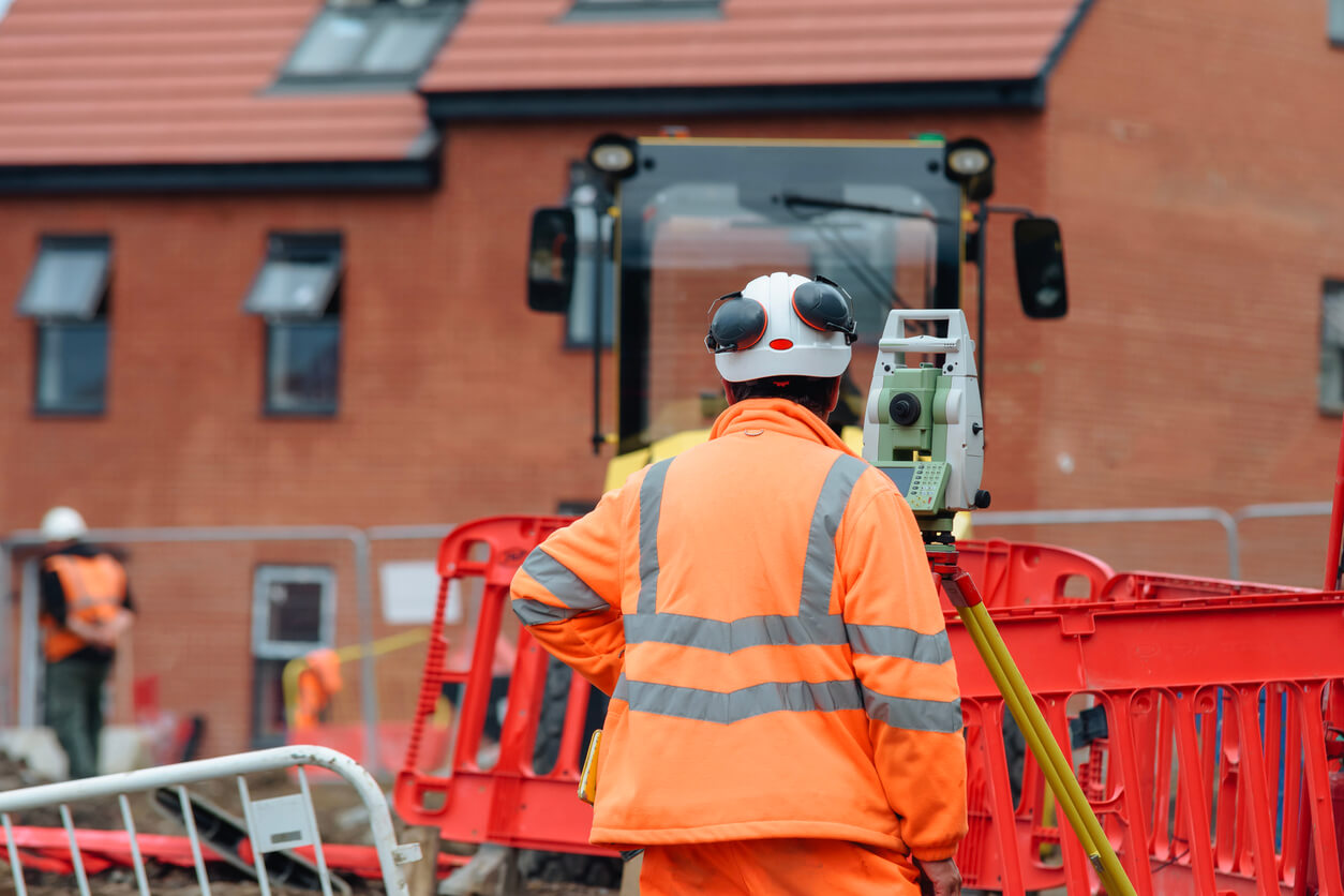 Construction worker in bright orange safety gear is using surveying instrument at construction site bordered by safety barriers with nearby machinery and workers visible